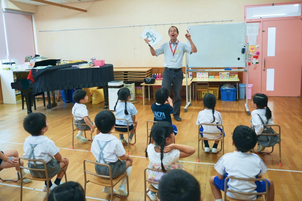 教室で絵を掲げる先生と、椅子に座って話を聞く子どもたち