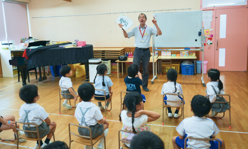 教室で絵を掲げる先生と、椅子に座って話を聞く子どもたち